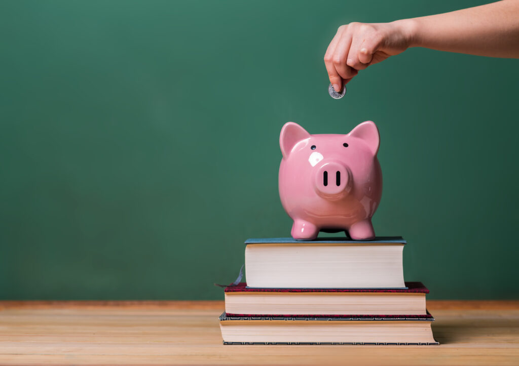 Person depositing money in a pink piggy bank on top of books with chalkboard in the background as concept image of the costs of education