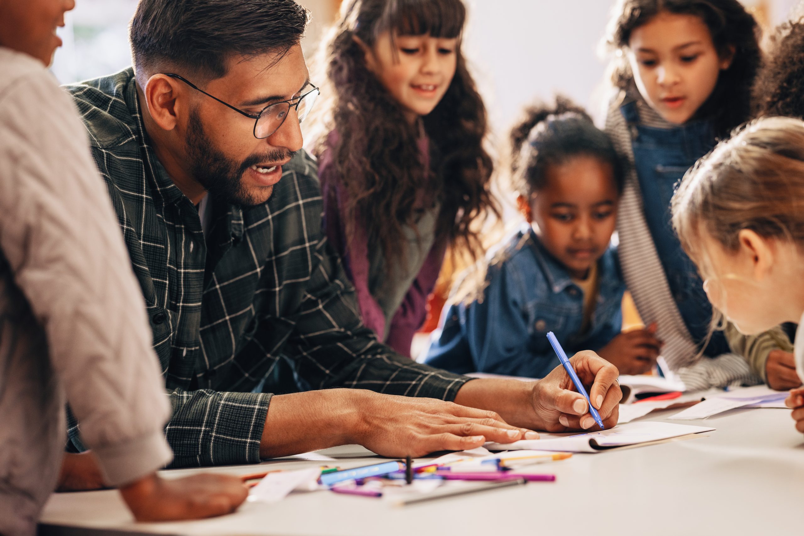 Man teaches his students how to draw in a primary school class. This elementary school educator shows his kids how to use a colouring pencil. Teaching an elementary class in a multiethnic school.