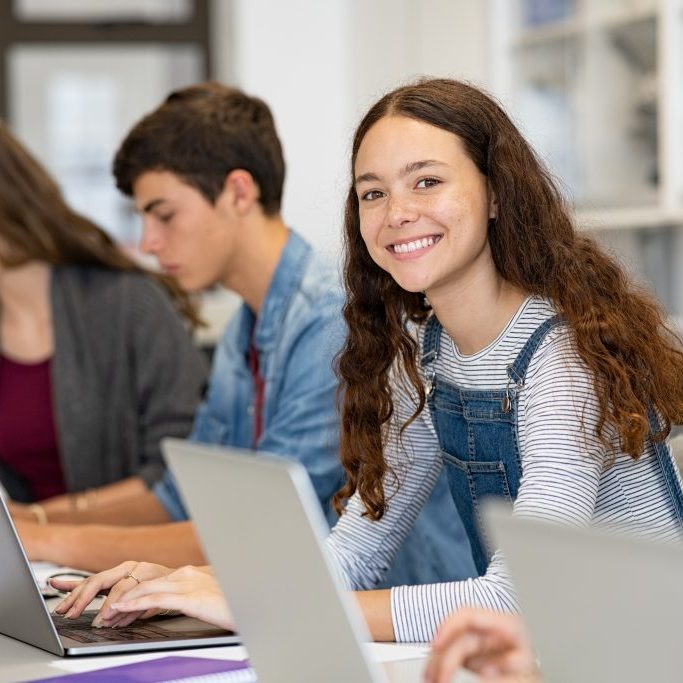 Happy young woman working on laptop and looking at camera in classroom. Portrait of smiling university student in library use computer for a research. Satisfied college student looking at camera while sitting in a row with classmates studying together.