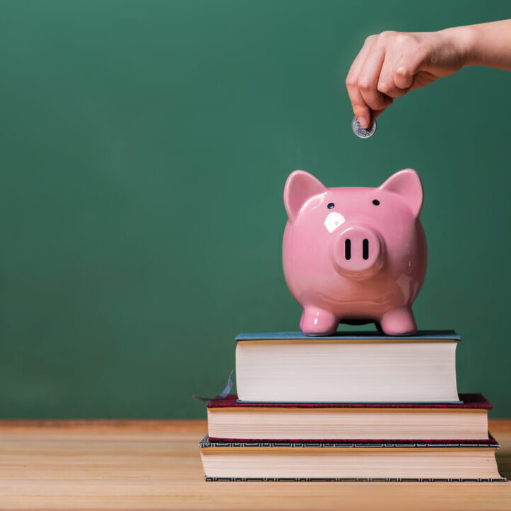 Person depositing money in a pink piggy bank on top of books with chalkboard in the background as concept image of the costs of education